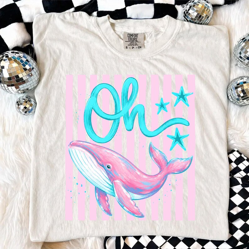 May include: Cream-colored t-shirt with a graphic featuring a pink whale, blue stars, and the word "Oh" in turquoise script. The design is set against a backdrop of pink vertical stripes.