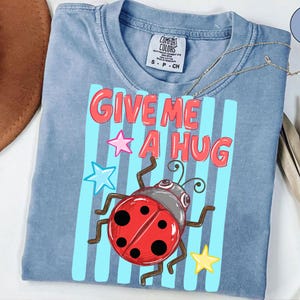 Pu&ograve; includere: T-shirt Comfort Colors azzurra con la scritta "GIVE ME A HUG" in rosso, bordata di bianco. Una coccinella dei cartoni animati e stelle colorate decorano la maglietta. La maglietta ha uno scollo tondo.