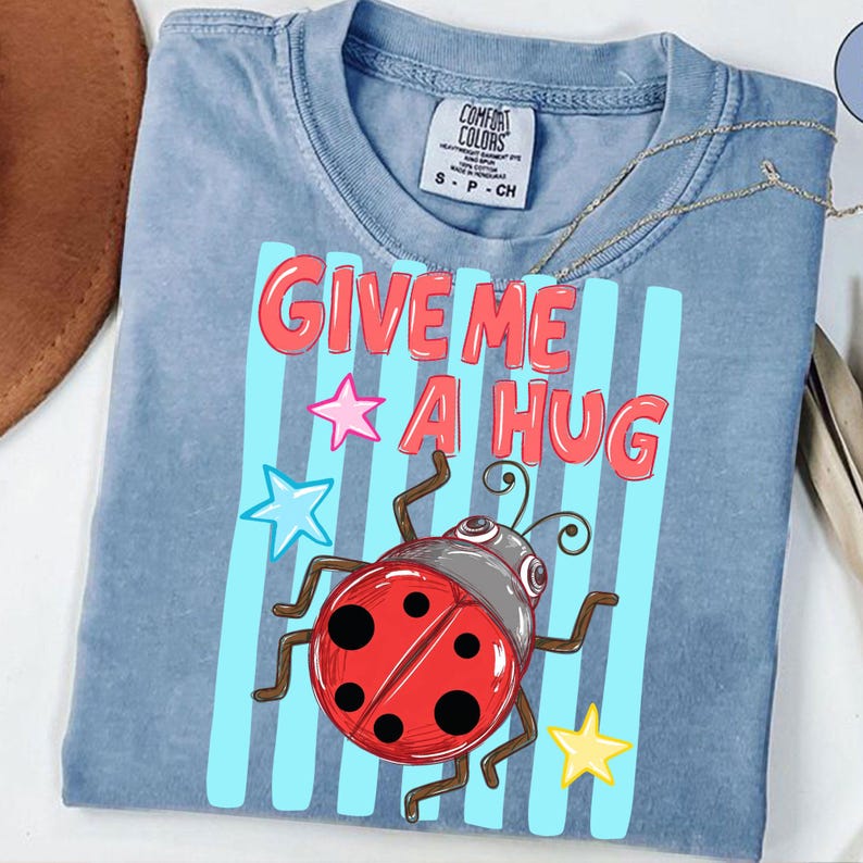 Pu&ograve; includere: T-shirt Comfort Colors azzurra con la scritta "GIVE ME A HUG" in rosso, bordata di bianco. Una coccinella dei cartoni animati e stelle colorate decorano la maglietta. La maglietta ha uno scollo tondo.