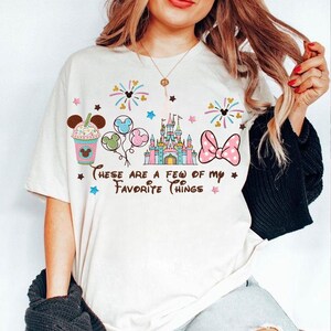 May include: Off-white t-shirt with a colorful graphic design featuring a castle, balloons, a bow, and a drink. The text "These are a few of my favorite things" is printed below the graphics. The shirt has short sleeves.