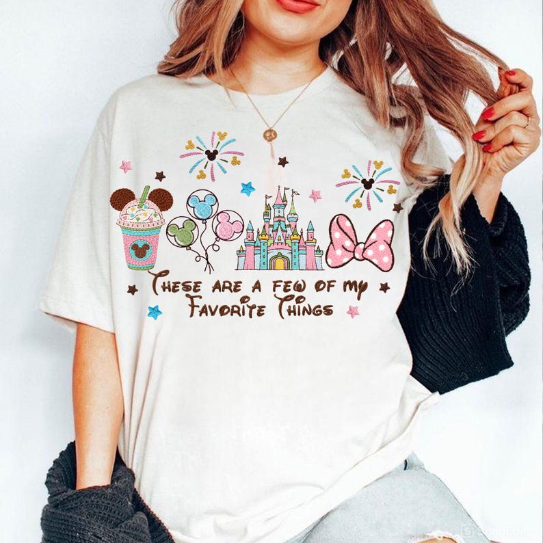 May include: Off-white t-shirt with a colorful graphic design featuring a castle, balloons, a bow, and a drink. The text "These are a few of my favorite things" is printed below the graphics. The shirt has short sleeves.