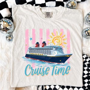 May include: Cream-coloured t-shirt featuring a colourful cruise ship design with the text "Cruise Time." The graphic includes a sun, pink stripes, and stars. The shirt is a Comfort Colors brand.