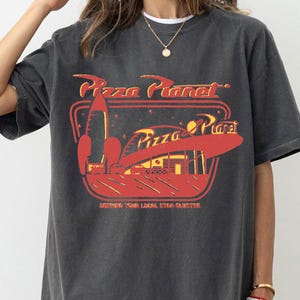 Puede incluir: Camiseta gris oscuro con un dise&ntilde;o rojo y amarillo de Pizza Planet, con un cohete y el texto "Serving Your Local Star Cluster". La camiseta tiene un aspecto vintage.