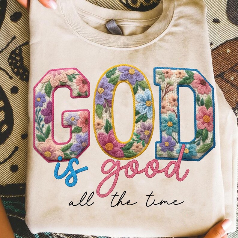 Op de afbeelding: Cr&egrave;mekleurig T-shirt met de tekst "GOD is good all the time". De letters van "GOD" zijn gevuld met bloemenborduurwerk in roze, paars en blauw. Het woord "good" is cursief geschreven.