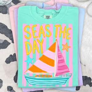 May include: A stack of Comfort Colors t-shirts in pastel colors. The top shirt is mint green with the words "SEAS THE DAY" in yellow, and a sailboat graphic. The sailboat has an orange and pink striped sail, and a blue hull.