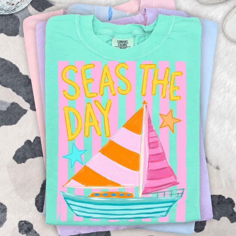 May include: A stack of Comfort Colors t-shirts in pastel colors. The top shirt is mint green with the words "SEAS THE DAY" in yellow, and a sailboat graphic. The sailboat has an orange and pink striped sail, and a blue hull.