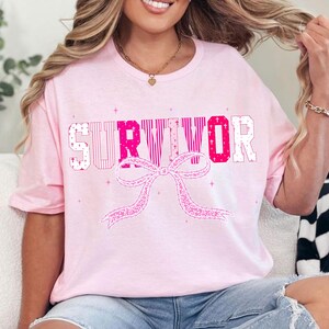 May include: A light pink t-shirt with the word "SURVIVOR" in a mix of pink and white block letters. A pink ribbon graphic is below the text. The shirt has short sleeves.