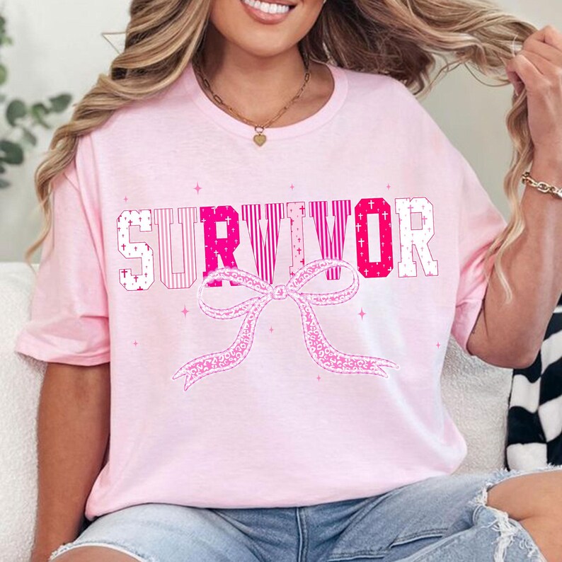 May include: A light pink t-shirt with the word "SURVIVOR" in a mix of pink and white block letters. A pink ribbon graphic is below the text. The shirt has short sleeves.