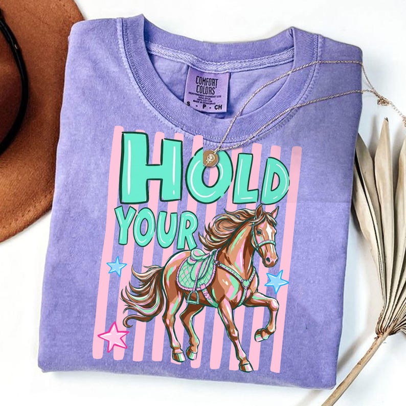 May include: A lilac Comfort Colors t-shirt featuring a horse illustration with the text "HOLD YOUR" in teal and pink. The horse is brown with a teal saddle and bridle. Pink and blue stars are scattered around the horse.