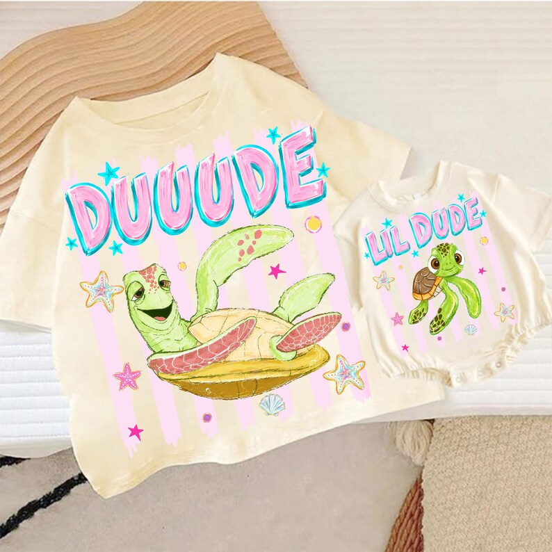 May include: Two matching cream-colored shirts. One is a t-shirt with the word "DUUUDE" in pink and blue, and a cartoon turtle. The other is a baby romper with "LIL DUDE" and a turtle graphic.