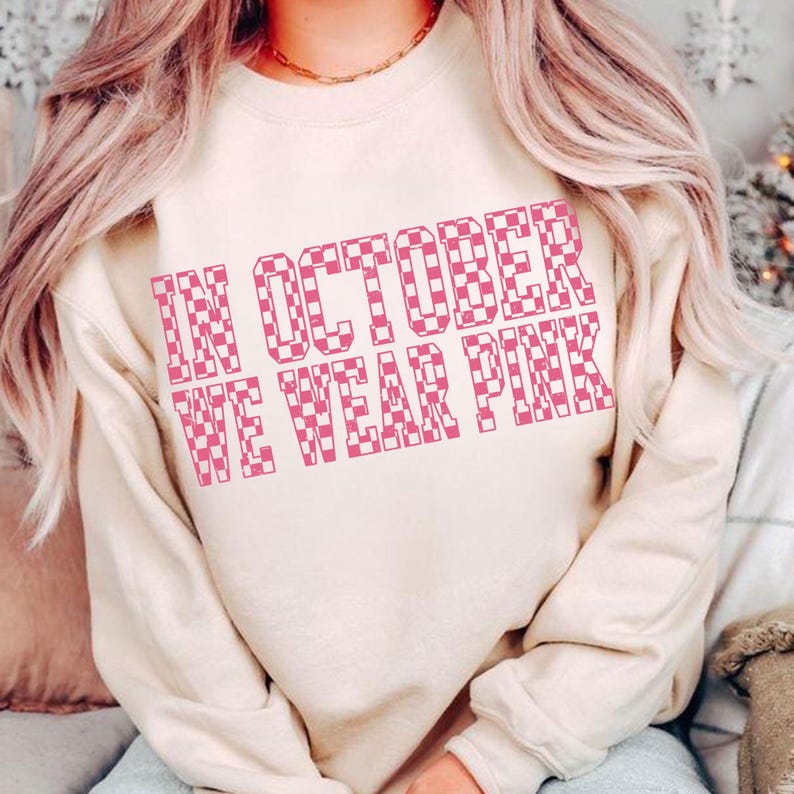May include: A beige sweatshirt with the text "IN OCTOBER WE WEAR PINK" in a pink checkered pattern. The sweatshirt has a crew neck and long sleeves. The text is centered on the front of the shirt.