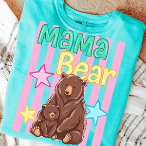 Pu&ograve; includere: Una maglietta turchese con le parole "Mama Bear" in verde e giallo, con strisce verticali rosa e stelle. Un orso bruno e il suo cucciolo sono raffigurati sotto il testo, creando un design toccante.