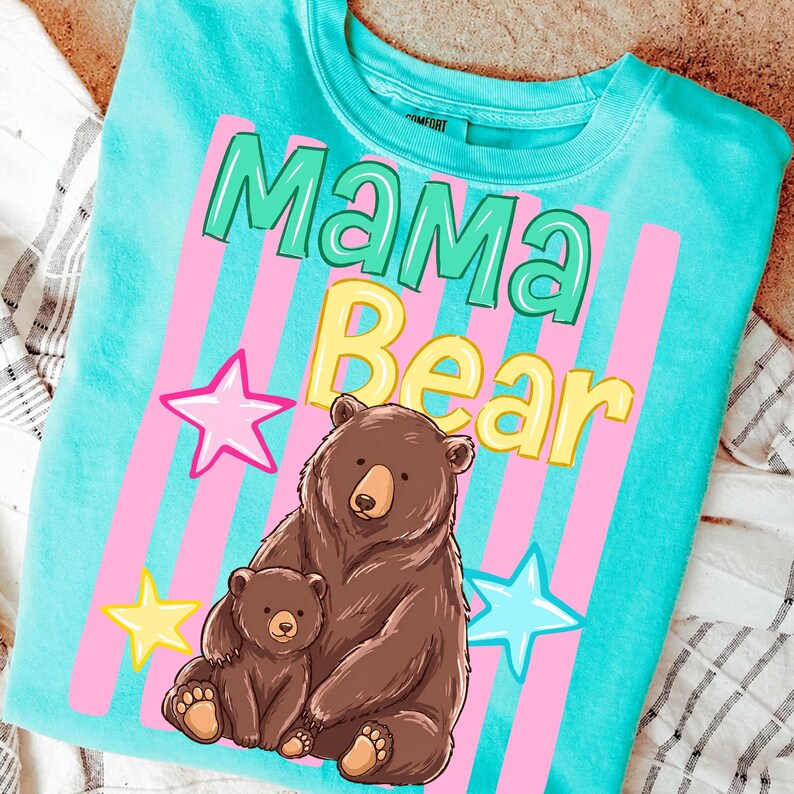 Pu&ograve; includere: Una maglietta turchese con le parole "Mama Bear" in verde e giallo, con strisce verticali rosa e stelle. Un orso bruno e il suo cucciolo sono raffigurati sotto il testo, creando un design toccante.