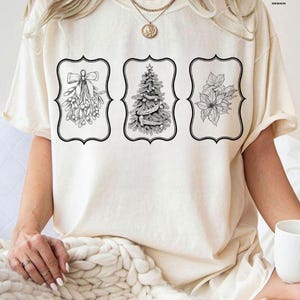 May include: Cream-colored t-shirt featuring three black and white framed illustrations. The illustrations include mistletoe, a Christmas tree, and poinsettias. The shirt has short sleeves and a relaxed fit, suitable for casual wear.