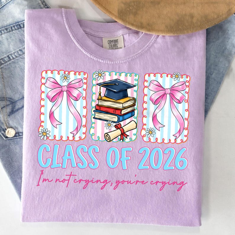 May include: A lilac Comfort Colors t-shirt with a graduation-themed design. The design includes pink bows, a graduation cap, books, and a scroll. The text reads "CLASS OF 2026" and "I'm not crying, you're crying."