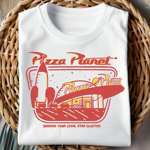 Puede incluir: Camiseta blanca con un gr&aacute;fico de Pizza Planet de estilo retro en rojo y amarillo. El dise&ntilde;o incluye un cohete, un edificio y el texto "Pizza Planet" y "Serving Your Local Star Cluster."