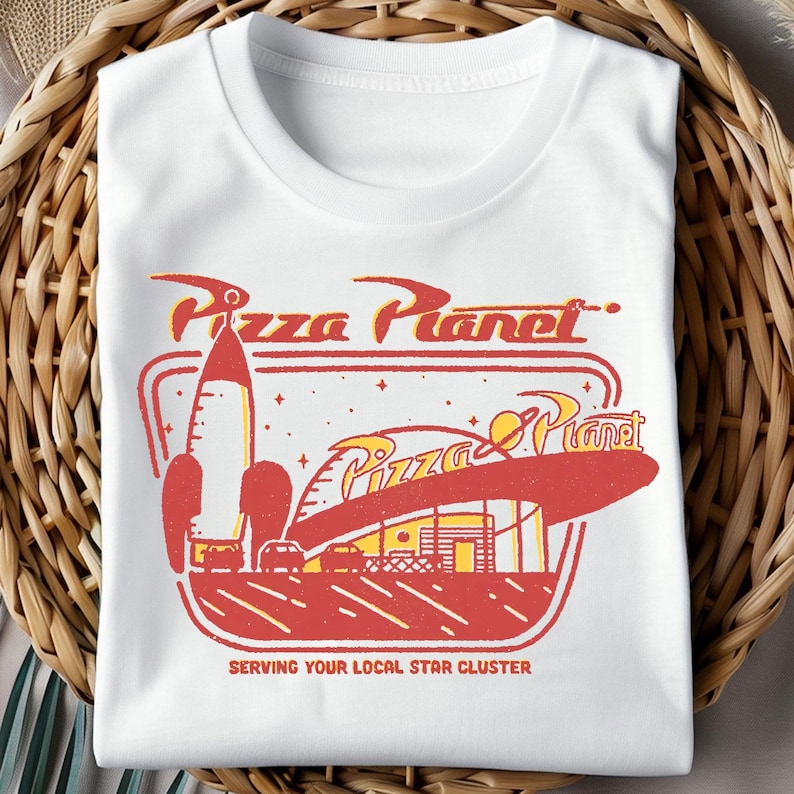 Puede incluir: Camiseta blanca con un gr&aacute;fico de Pizza Planet de estilo retro en rojo y amarillo. El dise&ntilde;o incluye un cohete, un edificio y el texto "Pizza Planet" y "Serving Your Local Star Cluster."