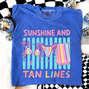Peut inclure: T-shirt bleu avec l'inscription "SUNSHINE AND TAN LINES" en rose et jaune. Le motif repr&eacute;sente un bikini et une serviette suspendus &agrave; une corde &agrave; linge. Le t-shirt est de la marque Comfort Colors, taille S-P-CH.