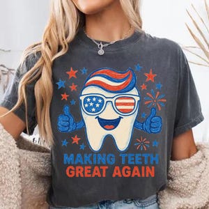 May include: Dark gray t-shirt with a patriotic tooth graphic. The tooth wears sunglasses with an American flag design and gives two thumbs up. The text "MAKING TEETH GREAT AGAIN" is printed below the tooth, surrounded by stars and fireworks.