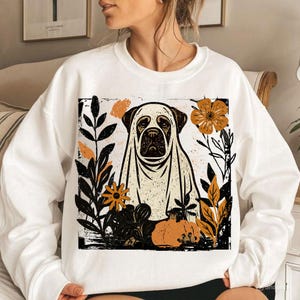 Pu&ograve; includere: Felpa bianca con grafica di un cane vestito da fantasma, circondato da foglie autunnali e una zucca. Il design include elementi neri, arancioni e beige, creando un tema stagionale. La felpa &egrave; un capo casual.