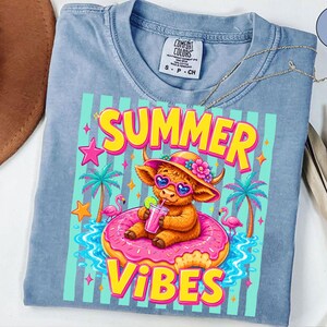 May include: Light blue Comfort Colors t-shirt with a colorful graphic. The graphic features a cartoon cow wearing sunglasses and a hat, sitting on a pink donut float with the words "SUMMER VIBES". Palm trees and flamingos are also in the design.