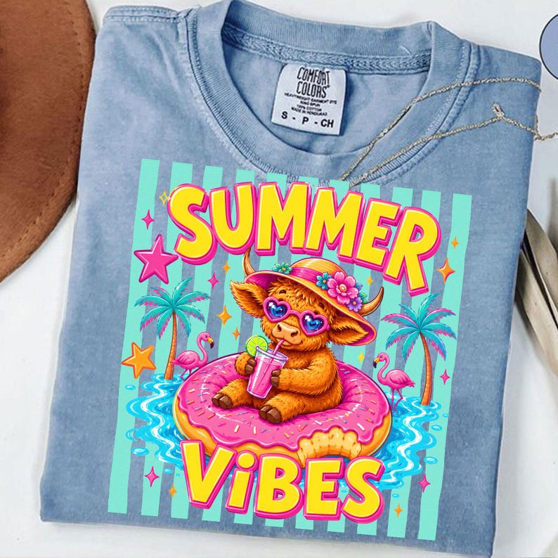 May include: Light blue Comfort Colors t-shirt with a colorful graphic. The graphic features a cartoon cow wearing sunglasses and a hat, sitting on a pink donut float with the words "SUMMER VIBES". Palm trees and flamingos are also in the design.
