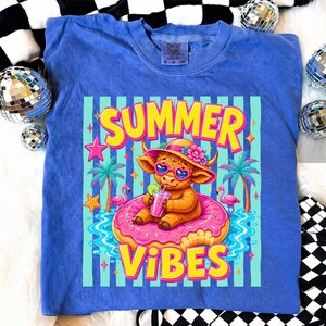 May include: Blue Comfort Colors t-shirt with a graphic of a Highland cow wearing sunglasses and a hat, floating on a pink donut in water. The text "SUMMER VIBES" is in yellow and pink.