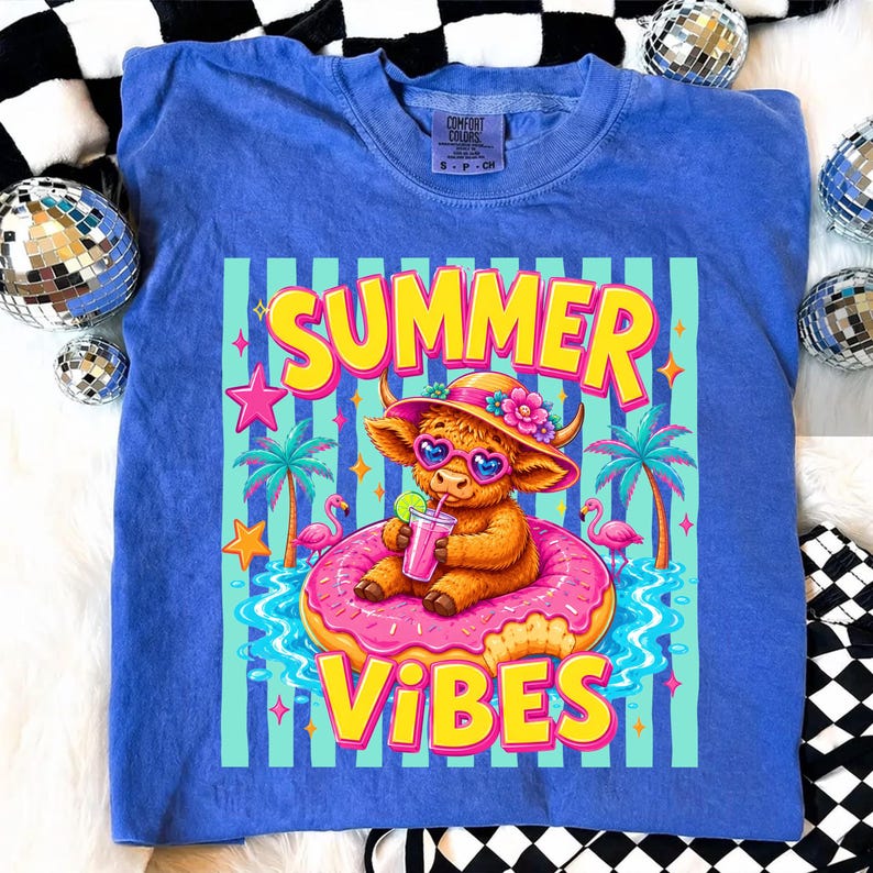 May include: Blue Comfort Colors t-shirt with a graphic of a Highland cow wearing sunglasses and a hat, floating on a pink donut in water. The text "SUMMER VIBES" is in yellow and pink.