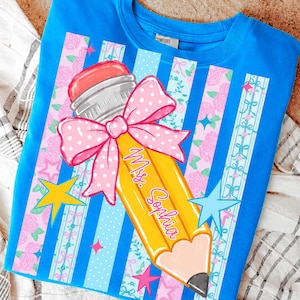 May include: A blue t-shirt with a large pencil graphic. The pencil is yellow with a pink eraser and a pink bow. The text "Mrs. Sophia" is written on the pencil. The shirt has a striped design with stars and floral accents.