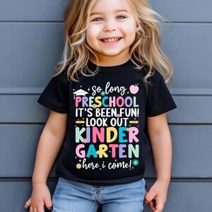 Puede incluir: Camiseta negra con texto colorido que dice "So long Preschool, It's Been Fun! Look Out Kindergarten, Here I Come!". La camiseta tiene gr&aacute;ficos de gorro de graduaci&oacute;n, manzana y flores.