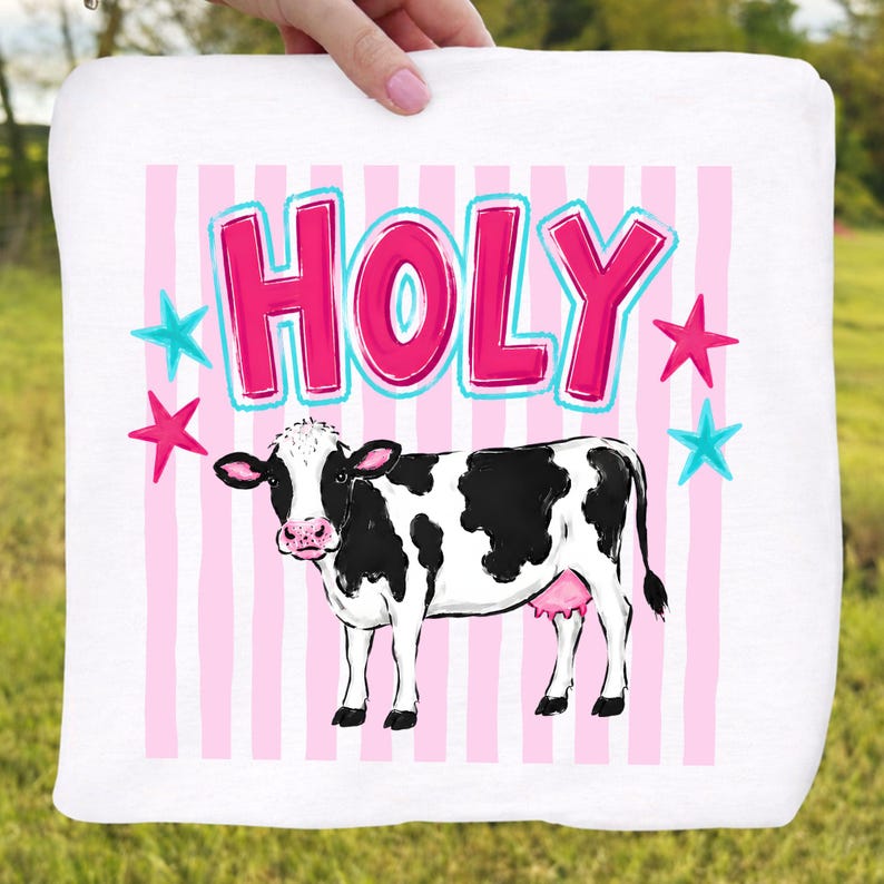 Puede incluir: Toalla blanca con una ilustraci&oacute;n de vaca de dibujos animados. La vaca es blanca y negra con una ubre rosa. La palabra "HOLY" est&aacute; escrita en letras rosas con un contorno azul, y hay estrellas rosas y azules.