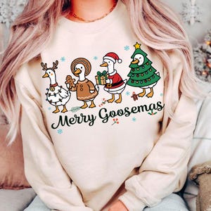Peut inclure: Un sweat-shirt beige avec un motif de No&euml;l. Il pr&eacute;sente quatre oies habill&eacute;es pour les f&ecirc;tes : une avec des bois et des lumi&egrave;res, une en pain d'&eacute;pices, une en P&egrave;re No&euml;l et une en sapin de No&euml;l. Le texte "Merry Goosemas" est en dessous.