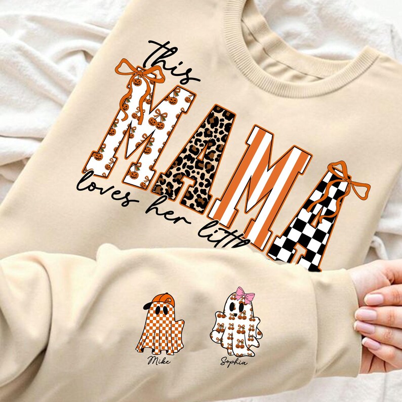 May include: Beige sweatshirt with the text "this MAMA loves her littles". The word "MAMA" is decorated with pumpkin and leopard print patterns. Two ghost illustrations are on the sleeve, one with a baseball cap and the other with a pink bow.