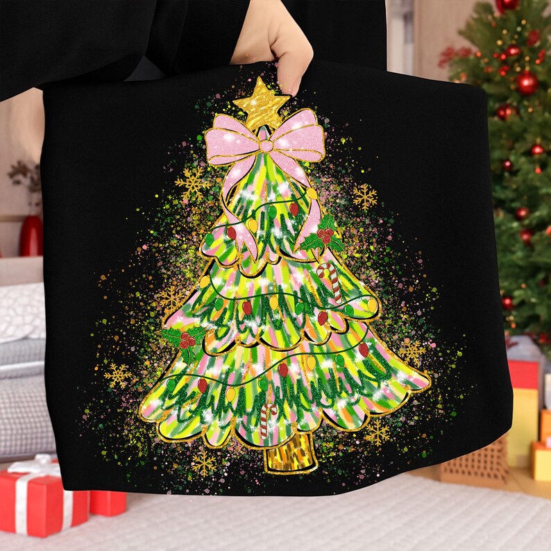 May include: Black tote bag featuring a colorful Christmas tree design. The tree is decorated with a pink bow, gold star, and holly berries. The background has a splatter effect with gold snowflakes.