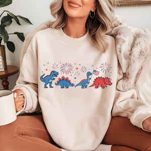 Puede incluir: Sudadera color crema con un dise&ntilde;o de dinosaurios de dibujos animados en azul y rojo, con fuegos artificiales y estrellas. Un dinosaurio sostiene una bandera americana. El dise&ntilde;o est&aacute; impreso en el pecho.