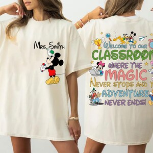 May include: Cream-colored t-shirt featuring Mickey Mouse holding a marker, with "Mrs. Smith" above. The back displays "Welcome to our Classroom" with Disney characters and the text "Where the Magic Never Stops and the Adventure Never Ends!"