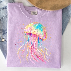 May include: A light purple t-shirt featuring a colorful watercolor jellyfish design. The jellyfish has a rainbow-colored cap and flowing tentacles in shades of blue, yellow, and pink. The shirt has a classic crew neck and short sleeves.