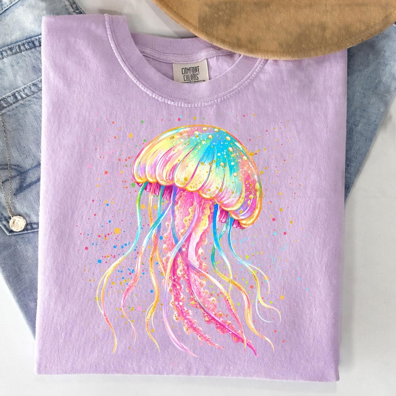 May include: A light purple t-shirt featuring a colorful watercolor jellyfish design. The jellyfish has a rainbow-colored cap and flowing tentacles in shades of blue, yellow, and pink. The shirt has a classic crew neck and short sleeves.