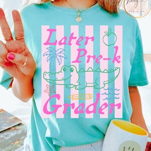 May include: A light blue t-shirt with a pink and white striped design. The shirt features the words "Later Pre-k Grader" in pink, along with a cartoon alligator, apple, palm tree, and other graphics.