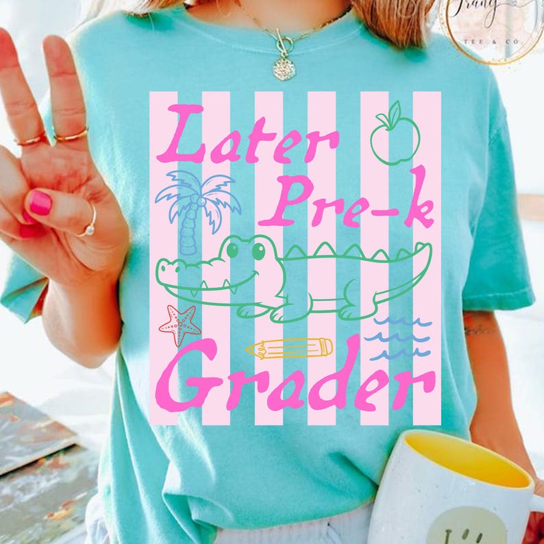 May include: A light blue t-shirt with a pink and white striped design. The shirt features the words "Later Pre-k Grader" in pink, along with a cartoon alligator, apple, palm tree, and other graphics.