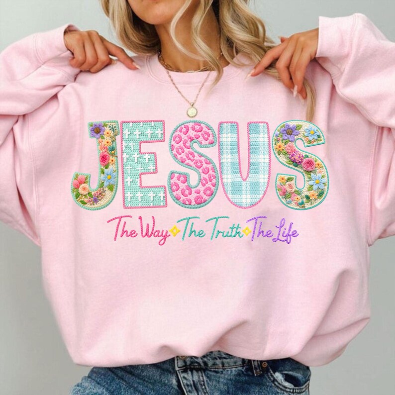 Puede incluir: Sudadera rosa claro con la palabra "JESUS" en letras grandes y coloridas. Las letras est&aacute;n decoradas con estampados florales, estampado de leopardo y un dise&ntilde;o a cuadros azul y blanco. Debajo de la palabra "JESUS" est&aacute; la frase "The Way The Truth The Life".