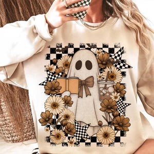 Könnte beinhalten: Beiges Sweatshirt mit einer Geisterillustration, die ein Buch und ein Getränk hält, umgeben von Blumen und Sternen. Das Design umfasst einen karierten Hintergrund und eine Fliege auf dem Geist. Ein lustiges, gruseliges Design.
