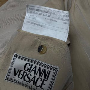 May include: Close-up of a tan-colored Gianni Versace Couture blazer. The interior lining is visible, showcasing a label with the brand's name and logo. A tag with product information is tucked into the pocket. The blazer has a button closure.