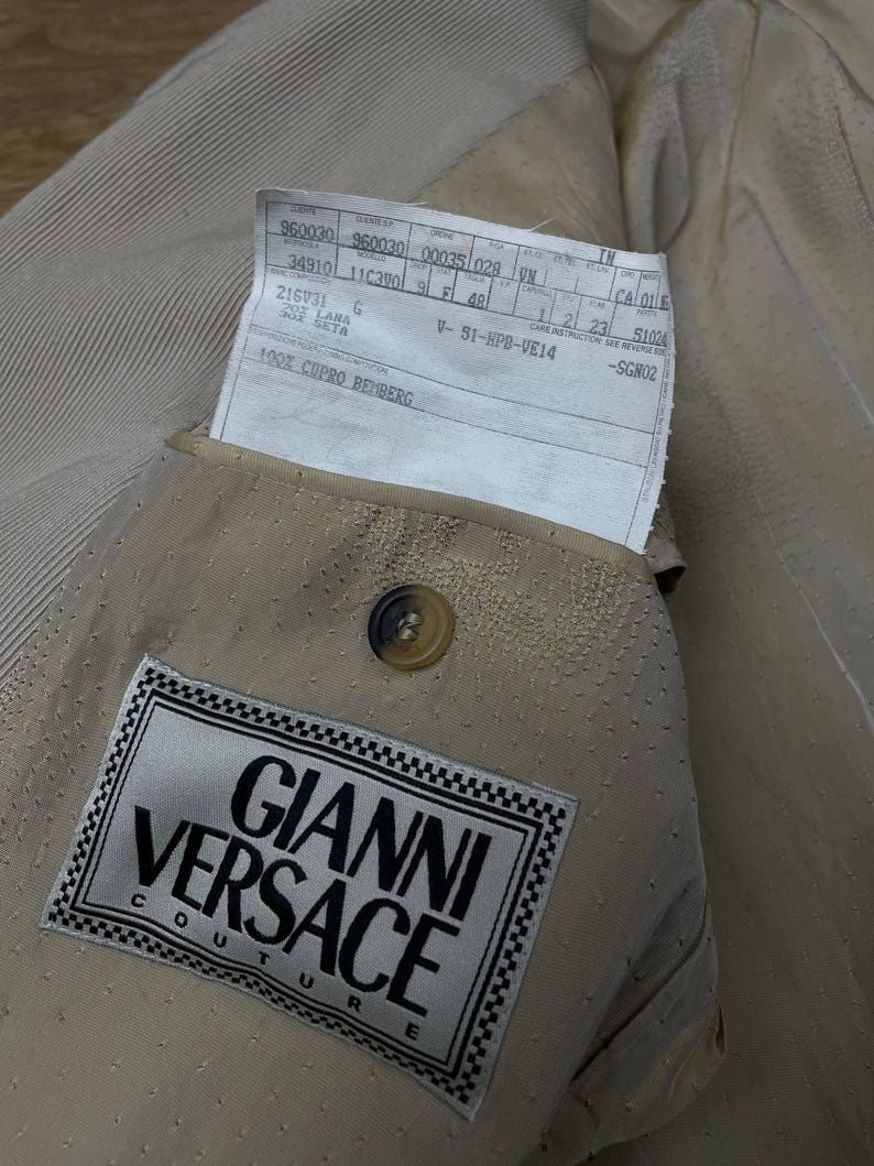 May include: Close-up of a tan-colored Gianni Versace Couture blazer. The interior lining is visible, showcasing a label with the brand's name and logo. A tag with product information is tucked into the pocket. The blazer has a button closure.