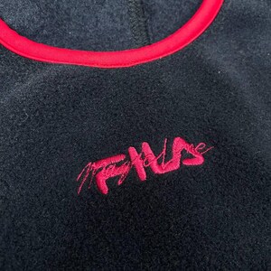 May include: A black fleece pullover featuring a vibrant red trim around the neckline. The garment displays the word FILA embroidered in a stylised, cursive red font on the chest, contrasting against the dark, textured fabric of the fleece material.