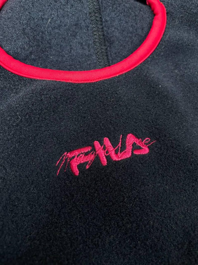 May include: A black fleece pullover featuring a vibrant red trim around the neckline. The garment displays the word FILA embroidered in a stylised, cursive red font on the chest, contrasting against the dark, textured fabric of the fleece material.
