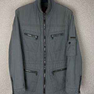 Può includere: Una giacca utility verde salvia con colletto alto foderato in pelliccia sintetica, chiusura frontale con zip integrale e quattro tasche orizzontali con zip sul petto e in vita. Le maniche presentano polsini con zip e una piccola tasca.