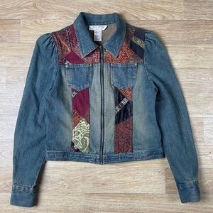 Puede incluir: Chaqueta vaquera azul con detalles de patchwork. La chaqueta tiene cuello, cremallera y dos bolsillos delanteros. El patchwork incluye estampados de cachemira y florales en tonos rojos, morados y dorados.