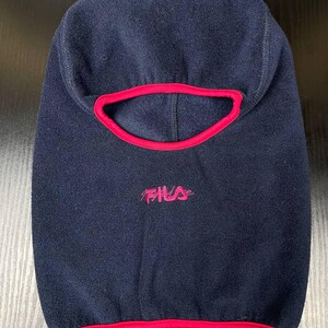 May include: A navy blue fleece balaclava featuring a bright pink trim around the face opening and the bottom hem. The word Fila is embroidered in pink thread on the front centre of the headgear, positioned below the eye opening.