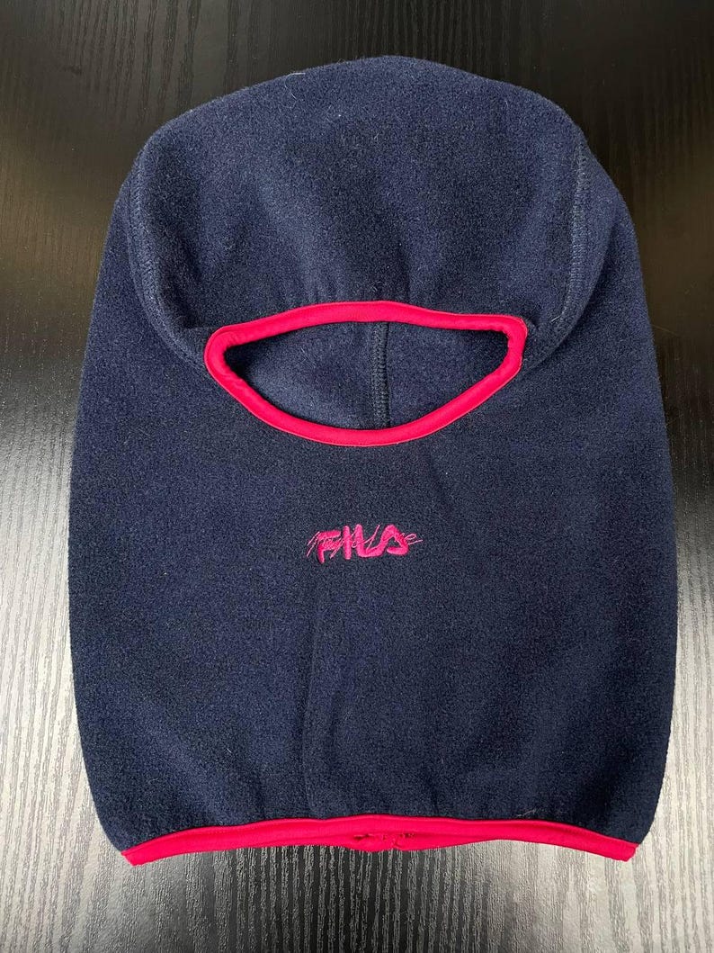 May include: A navy blue fleece balaclava featuring a bright pink trim around the face opening and the bottom hem. The word Fila is embroidered in pink thread on the front centre of the headgear, positioned below the eye opening.